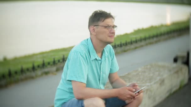 Молодой счастливый человек со смартфоном в городском парке сидит на берегу — стоковое видео