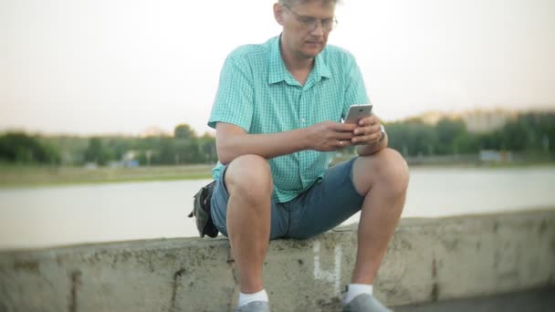 Joven hombre feliz con un teléfono inteligente en el parque de la ciudad se sienta en la orilla — Vídeos de Stock