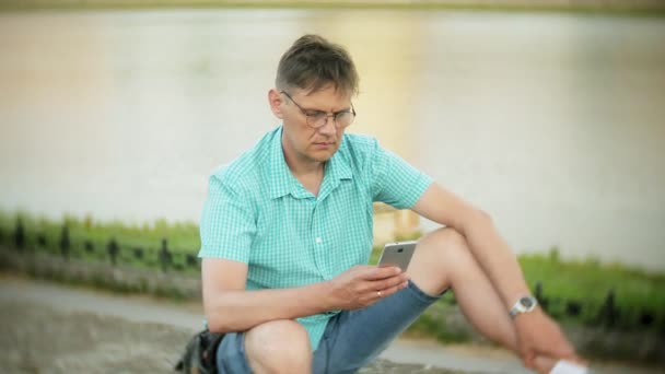 Joven hombre feliz con un teléfono inteligente en el parque de la ciudad se sienta en la orilla — Vídeos de Stock