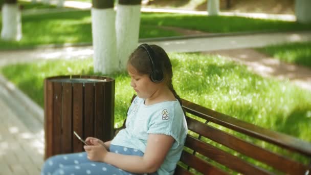 Niña gorda con una tablet PC y auriculares sentados en un banco escuchando música o viendo un video en un parque de verano — Vídeos de Stock