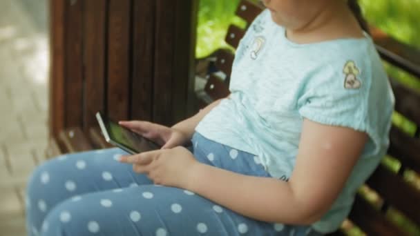 Petite fille grosse avec une tablette PC et écouteurs assis sur un banc à écouter de la musique ou regarder une vidéo dans un parc d'été — Video
