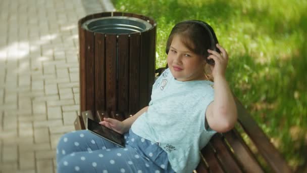 Bambina grassa con tablet PC e cuffie seduta su una panchina ad ascoltare musica o guardare un video in un parco estivo — Video Stock