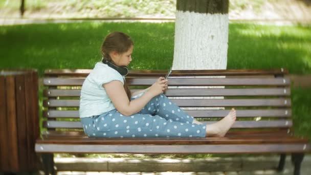 Маленькая толстая девочка с планшетным компьютером и наушниками, сидящая на скамейке и слушающая музыку или смотрящая видео в летнем парке — стоковое видео