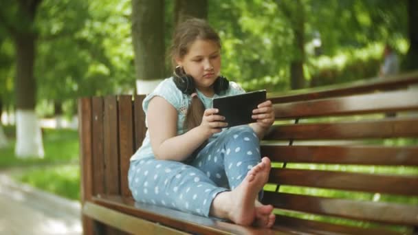 Маленька товста дівчинка з планшетним ПК і навушниками сидить на лавці, слухаючи музику або переглядаючи відео в літньому парку — стокове відео