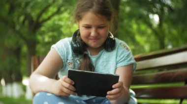 Küçük bir tablet Pc ile şişman kız ve müzik gerekse video bir yaz parkta bir bankta oturan kulaklıklar