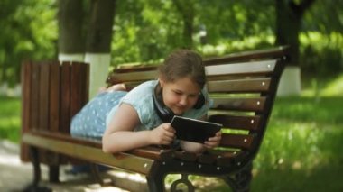 Küçük bir tablet Pc ile şişman kız ve müzik gerekse video bir yaz parkta bir bankta oturan kulaklıklar