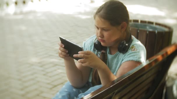 Niña gorda con una tablet PC y auriculares sentados en un banco escuchando música o viendo un video en un parque de verano — Vídeos de Stock