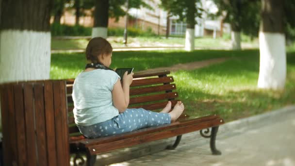Little fat girl with a tablet PC and headphones sitting on a bench listening to music or watching a video in a summer park — Stock Video