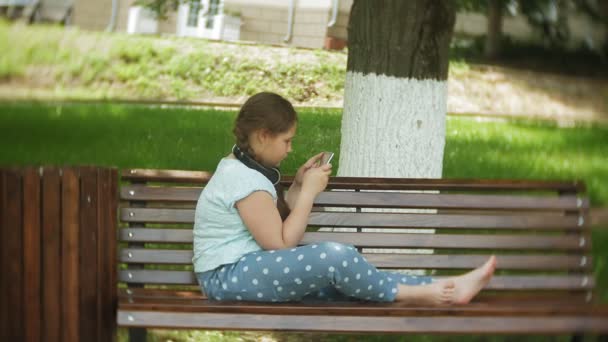 Маленька товста дівчинка з планшетним ПК і навушниками сидить на лавці, слухаючи музику або переглядаючи відео в літньому парку — стокове відео