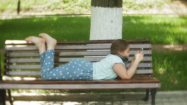 Little fat girl with a tablet PC and headphones sitting on a bench listening to music or watching a video in a summer park — Stock Video