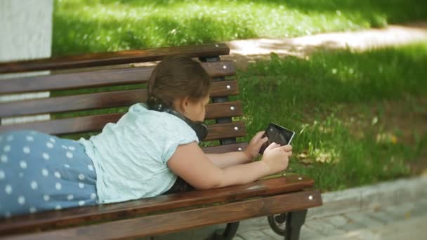小胖女孩与平板电脑和耳机坐在长凳上听音乐或观看视频在一个夏季公园 — 图库视频影像