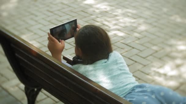 Lite fett flicka med en tablet Pc och hörlurar som sitter på en bänk lyssnar på musik eller titta på en video i en sommar park — Stockvideo