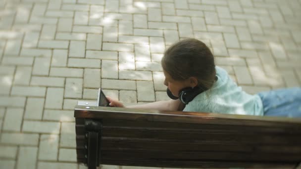 Niña gorda con una tablet PC y auriculares sentados en un banco escuchando música o viendo un video en un parque de verano — Vídeo de stock