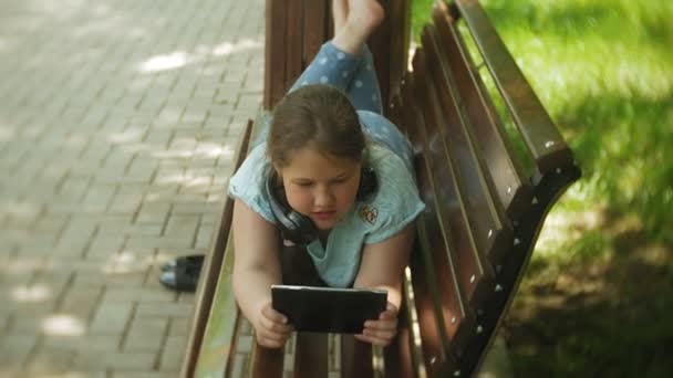 Petite fille grosse avec une tablette PC et écouteurs assis sur un banc à écouter de la musique ou regarder une vidéo dans un parc d'été — Video