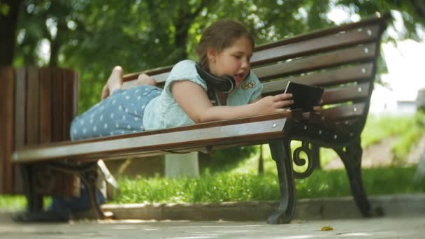 Niña gorda con una tablet PC y auriculares sentados en un banco escuchando música o viendo un video en un parque de verano — Vídeo de stock