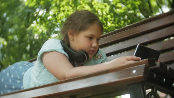 Niña gorda con una tablet PC y auriculares sentados en un banco escuchando música o viendo un video en un parque de verano — Vídeos de Stock