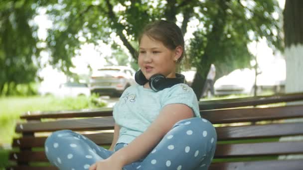 Niña gorda con una tablet PC y auriculares sentados en un banco escuchando música o viendo un video en un parque de verano — Vídeo de stock