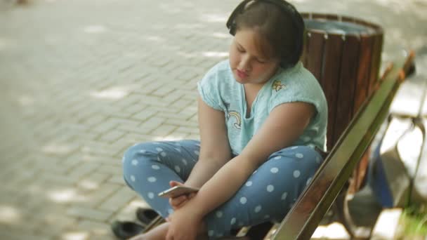 Niña gorda con una tablet PC y auriculares sentados en un banco escuchando música o viendo un video en un parque de verano — Vídeo de stock