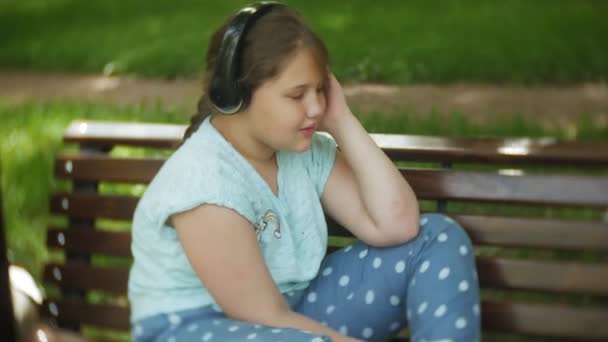 Niña gorda con una tablet PC y auriculares sentados en un banco escuchando música o viendo un video en un parque de verano — Vídeos de Stock