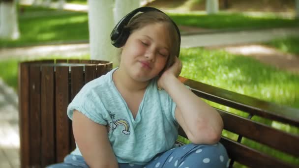 Niña gorda con una tablet PC y auriculares sentados en un banco escuchando música o viendo un video en un parque de verano — Vídeos de Stock