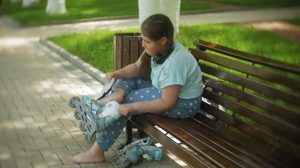 Λίγο λίπος κορίτσι που κάθεται σε ένα παγκάκι ακούγοντας μουσική ρούχα πατίνια στο πάρκο — Αρχείο Βίντεο