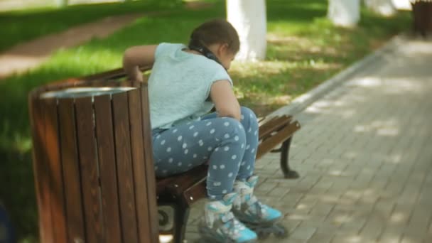 Lite fett flicka sitter på en bänk som lyssnar på musik kläder rullskridskor i parken — Stockvideo