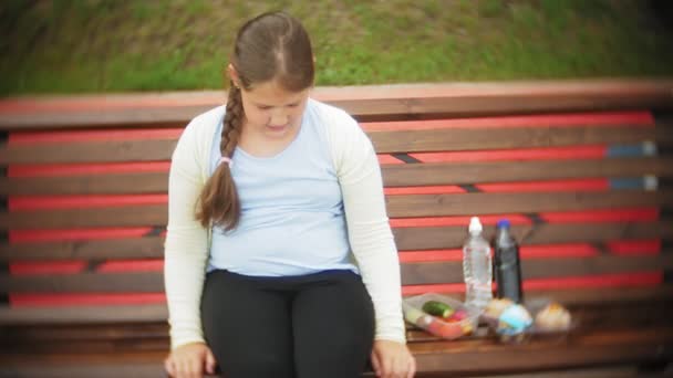 Крупный план очаровательной маленькой толстой девочки, ныряющей между тортом и овощами, сидящей на скамейке в кафе, концепция здоровой диеты — стоковое видео