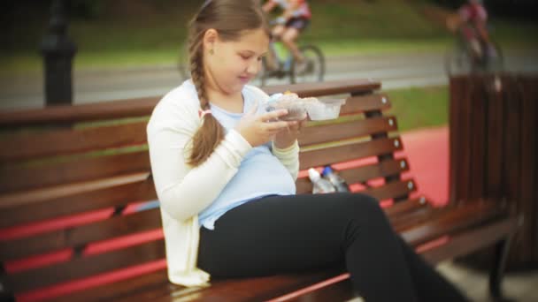 Крупным планом очаровательная маленькая девочка ест торт с руками, сидя на скамейке в парке, концепция здоровой диеты ... — стоковое видео