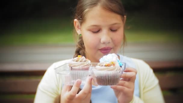 Ένα αξιολάτρευτο κοριτσάκι που τρώει ένα κέικ με τα χέρια της να κάθεται σε ένα παγκάκι στο πάρκο, η έννοια της υγιεινής διατροφής σε κοντινό πλάνο... — Αρχείο Βίντεο