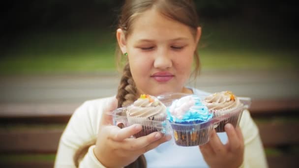 Nahaufnahme eines entzückenden kleinen Mädchens, das mit den Händen auf einer Bank im Park sitzt und einen Kuchen isst, das Konzept einer gesunden Ernährung... — Stockvideo