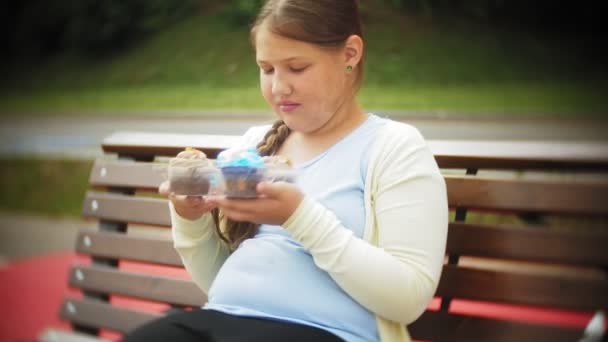 Primer plano de una adorable niña comiendo un pastel con las manos sentadas en un banco en el parque, el concepto de una dieta saludable ... — Vídeo de stock