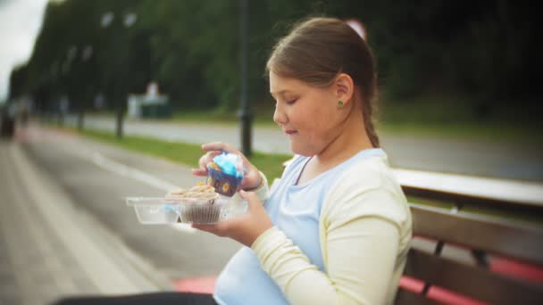 Close up de adorável menina gorda comendo um bolo com as mãos sentadas em um banco no parque — Vídeo de Stock