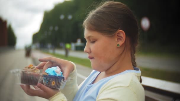 Nahaufnahme eines entzückenden kleinen dicken Mädchens, das mit den Händen auf einer Bank im Park sitzt und einen Kuchen isst — Stockvideo