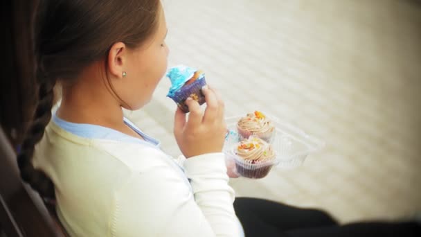 Close up de adorável menina gorda comendo um bolo com as mãos sentadas em um banco no parque — Vídeo de Stock