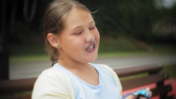 Close up de adorável menina gorda comendo um bolo com as mãos sentadas em um banco no parque — Vídeo de Stock