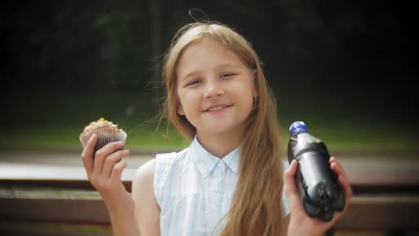 Nahaufnahme eines entzückenden kleinen Mädchens, das mit den Händen auf einer Bank im Park sitzt und einen Kuchen isst — Stockvideo