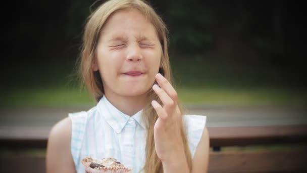 Gros plan d'une adorable petite fille mangeant un gâteau les mains assises sur un banc dans le parc — Video