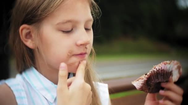 Close up de adorável menina comendo um bolo com as mãos sentadas em um banco no parque — Vídeo de Stock