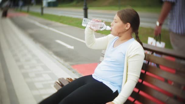 Маленька товста дівчинка на роликах п'є воду. Дитина п'є воду в парку на лавці — стокове відео