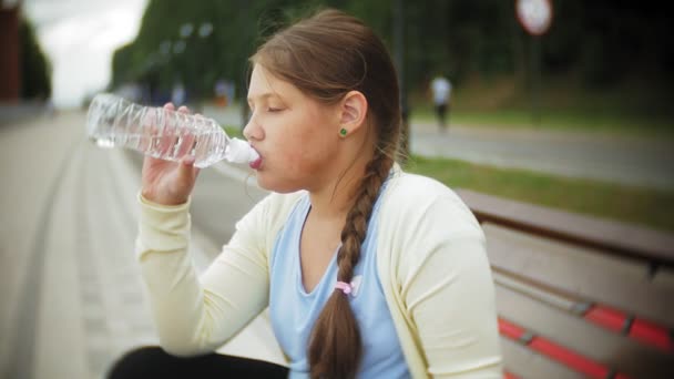 Una chica gorda sobre rodillos bebe agua. Un niño bebe agua en un parque en un banco — Vídeos de Stock