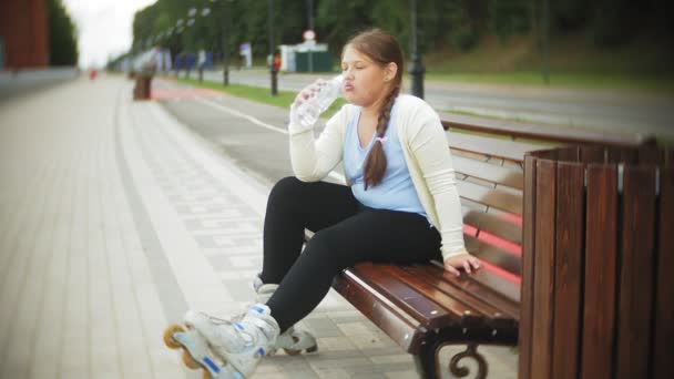 En liten fet tjej på rullarna dricker vatten. Ett barn dricker vatten i en park på en bänk — Stockvideo