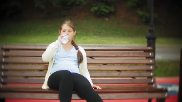 En liten fet tjej på rullarna dricker vatten. Ett barn dricker vatten i en park på en bänk — Stockvideo