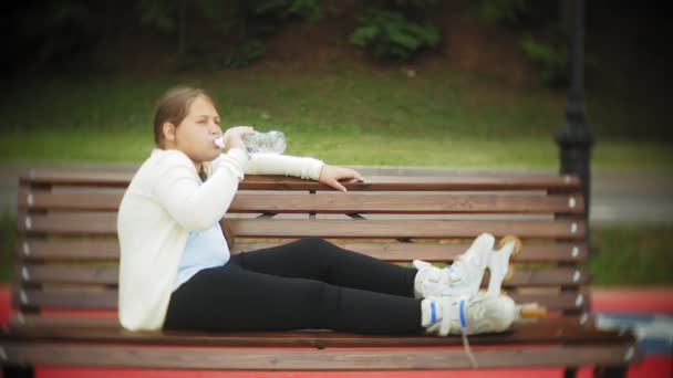 Uma pequena garota gorda em rolos bebe água. Uma criança bebe água em um parque em um banco — Vídeo de Stock