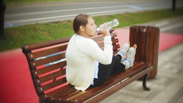 En liten fet tjej på rullarna dricker vatten. Ett barn dricker vatten i en park på en bänk — Stockvideo