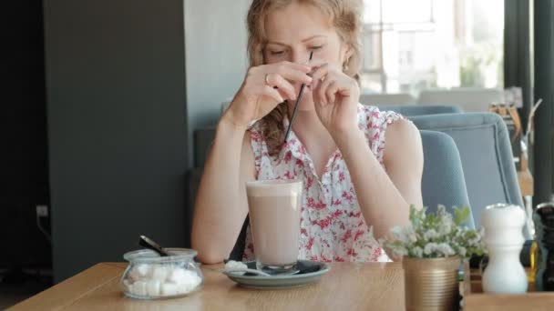 Счастливая девушка пьет кофе и отдыхает в кафе, улыбаясь и глядя на смартфон — стоковое видео