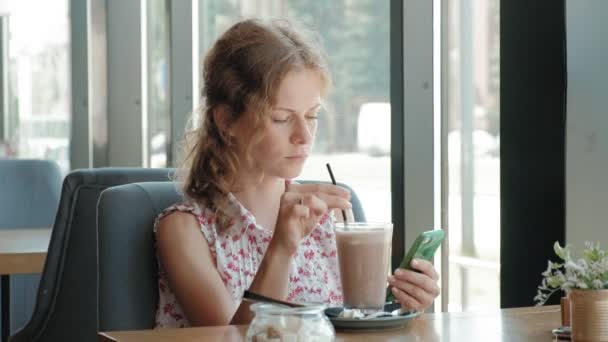 Šťastná dívka pití kávy a odpočinku v kavárně, s úsměvem a při pohledu na smartphone — Stock video