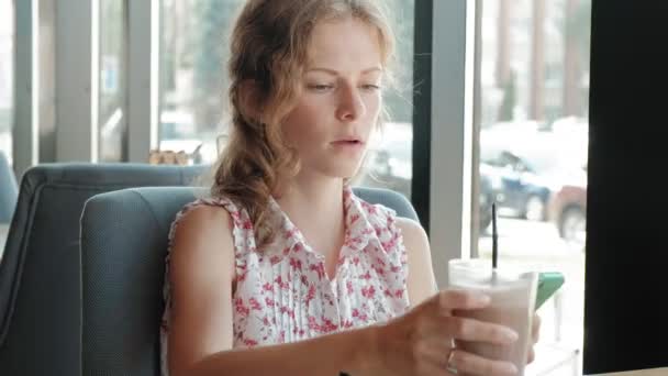 O fată fericită bea cafea și se relaxează în cafenea, zâmbind și uitându-se la smartphone — Videoclip de stoc