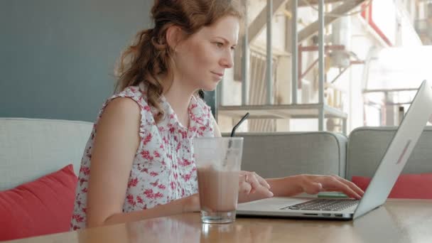 Una joven mujer de negocios se sienta en una mesa en un restaurante. Llevaba un portátil con acceso a Internet, revisa el correo. Se ve muy feliz. — Vídeos de Stock
