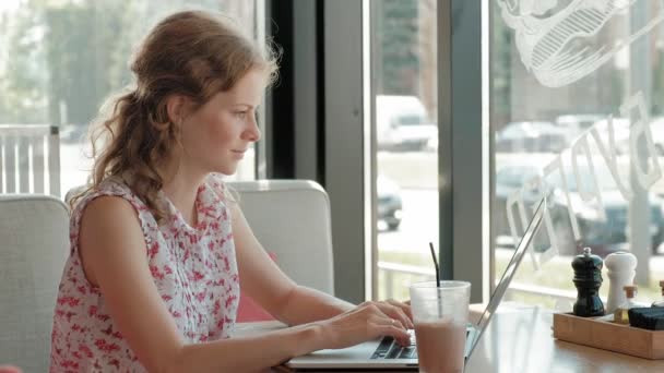 Jovem mulher de negócios senta-se em uma mesa em um restaurante. Ela carregava um portátil com acesso à Internet, ele verifica o correio. Ela parece muito feliz. — Vídeo de Stock
