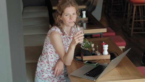 Jovem mulher de negócios senta-se em uma mesa em um restaurante. Ela carregava um laptop com acesso à Internet, verifica o correio e bebe café. Ela parece muito feliz. — Vídeo de Stock